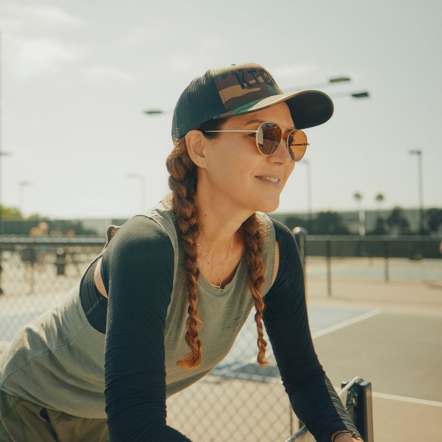 Woman with braided hair wearing a camo trucker hat and sunglasses outdoors.