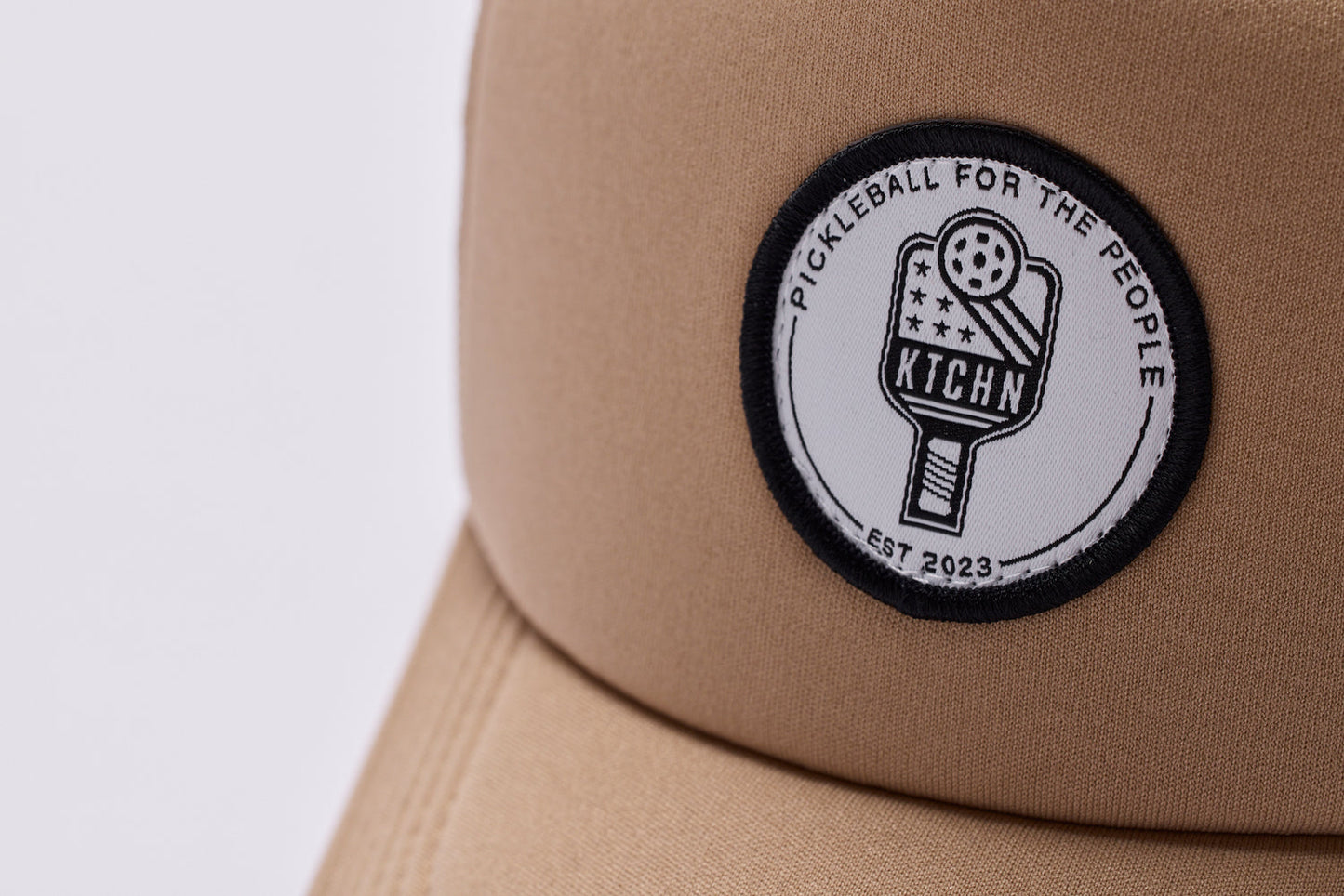 Tan foam trucker hat close-up, featuring a circular patch with a pickleball paddle design.