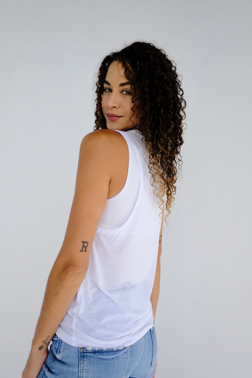 Woman in white muscle tank and jeans against a white background.