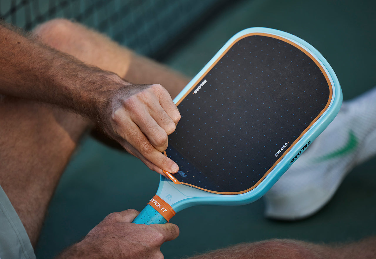 Applying replacement grip tape to a teal pickleball paddle outdoors.