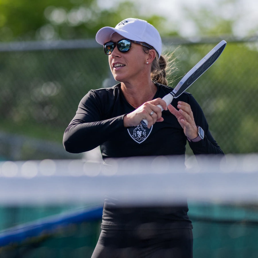 Woman playing pickleball outdoors, holding paddle, wearing sunglasses.