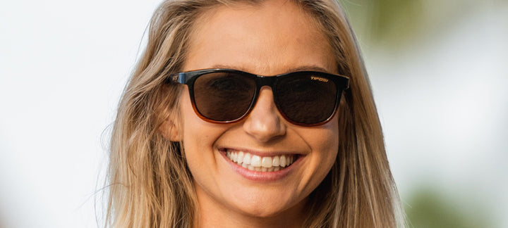 Woman smiling while wearing brown tortoiseshell sunglasses outdoors.