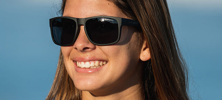 Woman smiling outdoors wearing matte black square sunglasses.