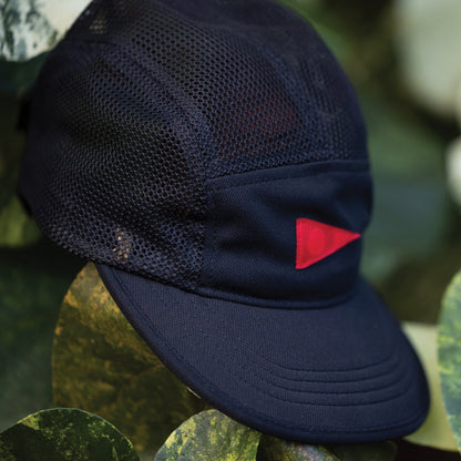 Black unstructured hat with breathable mesh panels and a small red detail, nestled amongst green foliage.
