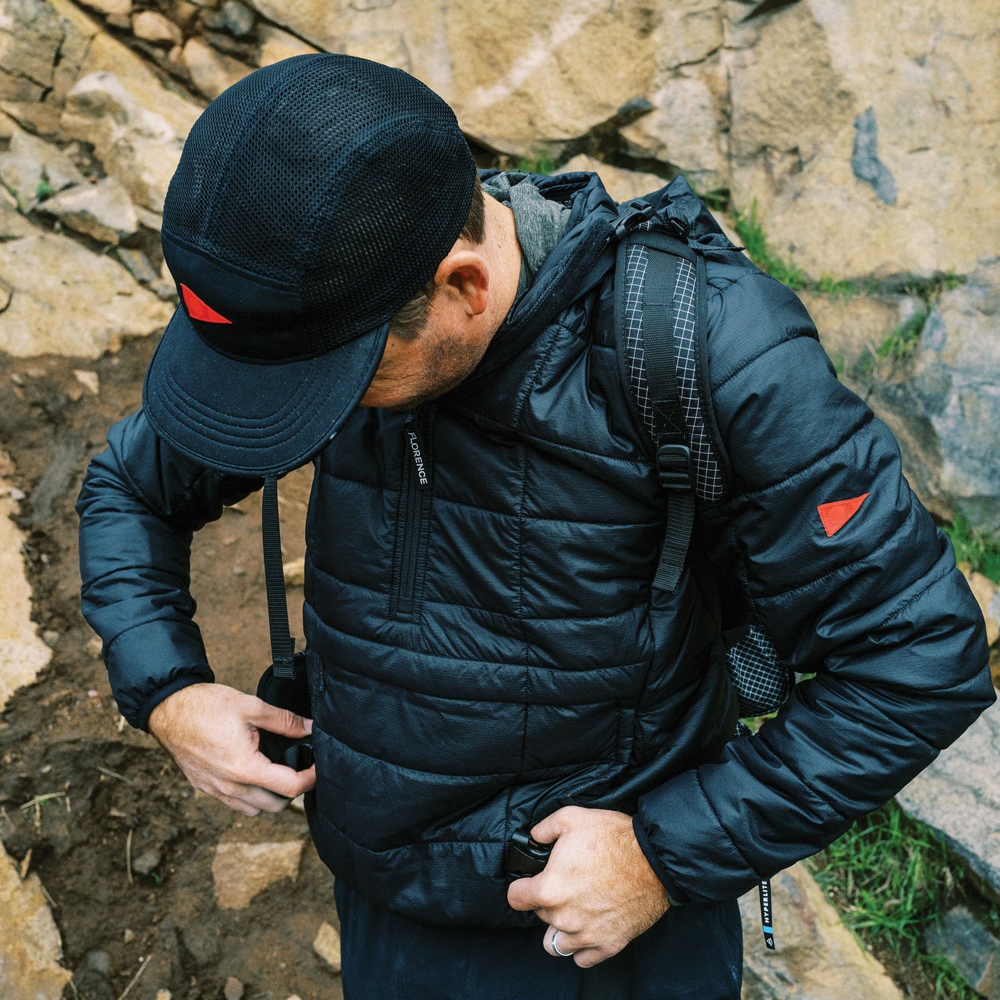 Man adjusting a dark puffer jacket outdoors near rocks.
