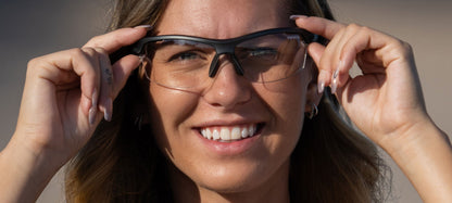 Woman adjusting matte gunmetal sunglasses with clear lenses outdoors.
