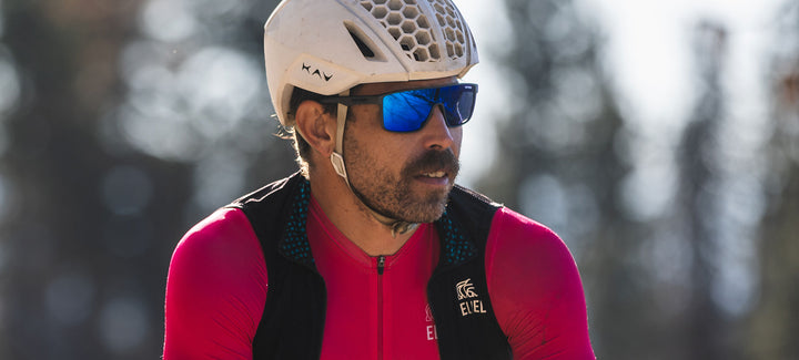 Cyclist wearing blue mirrored sunglasses and a pink jersey against a blurred outdoor background.
