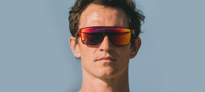 Man modeling red and purple sunglasses outdoors against a pale blue sky.
