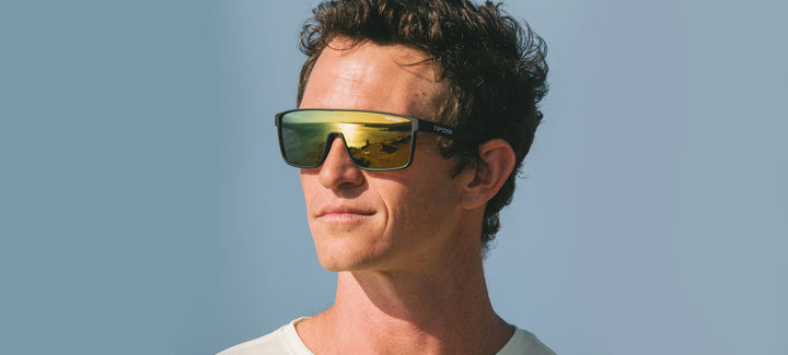 Man modeling matte black sunglasses with yellow-gold mirror lenses outdoors.
