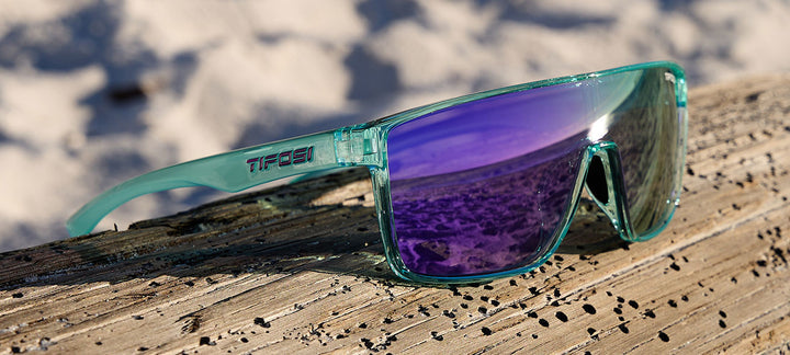 Aqua-colored sunglasses with purple mirrored lenses rest on weathered wood, beach in the background.
