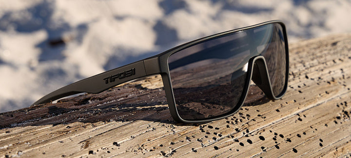 Black sunglasses resting on weathered wood, blurred beach background.
