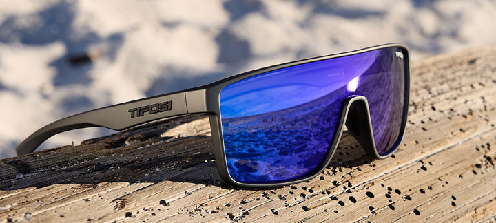 Matte gunmetal sunglasses with cobalt blue mirrored lenses, resting on weathered wood near a beach.
