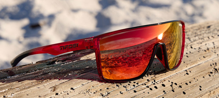 Red sunglasses with mirrored lenses, resting on weathered wood near a beach.
