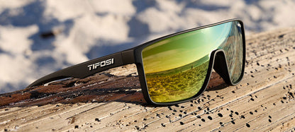 Matte black sunglasses with yellow-gold mirrored lenses rest on weathered wood, reflecting a beach scene.
