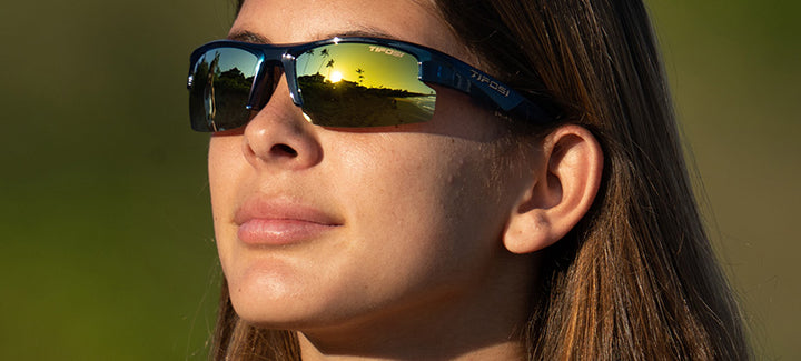 Woman wearing sporty sunglasses outdoors, reflecting sunset.
