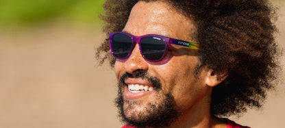 Man smiling outdoors wearing purple sunglasses with rainbow temples.
