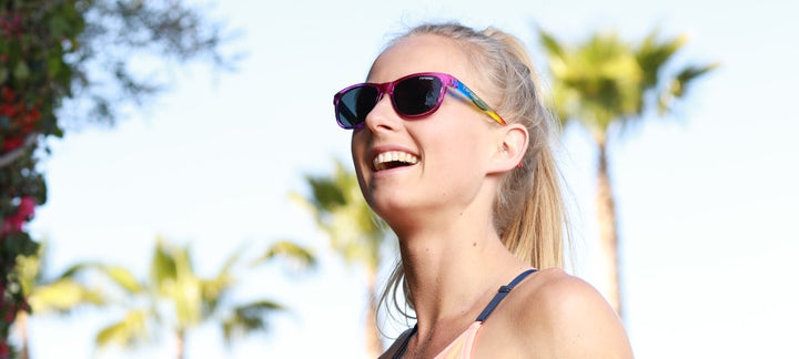 Woman smiling outdoors wearing vibrant sunglasses with a rainbow temple, enjoying sunny weather.
