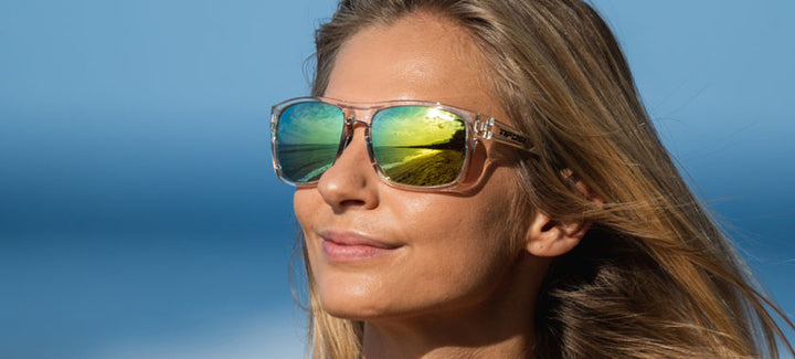 Clear frame sunglasses reflecting a sunny beach scene, worn by a woman outdoors.
