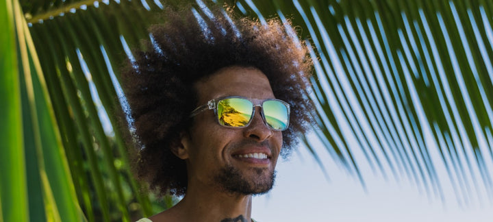 Man smiling outdoors, wearing clear-framed sunglasses with yellow-gold mirrored lenses, partially obscured by palm fronds.
